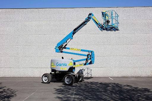 Een blauwe hoogtewerker met knikarm die omhoog staat van het merk Genie staat deels in de schaduw van een boom.